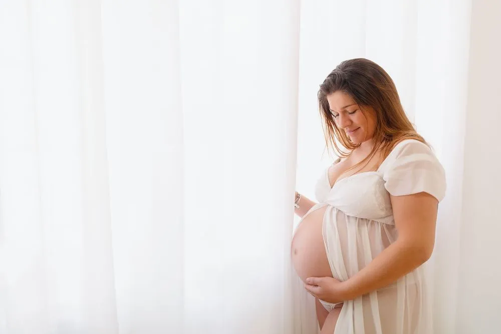 Grávida de vestido branco junto a cortinas claras, com as mãos na barriga