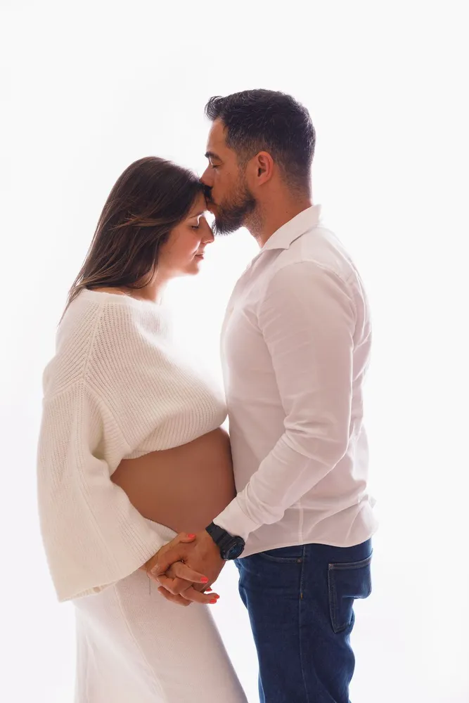 Casal no estúdio: ele beija-lhe a testa e as mãos de ambos estão sobre a barriga